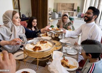 Middle Eastern woman in traditional attire smiling as she hands bearded man dish of roasted meat and creamy rice at midday meal.